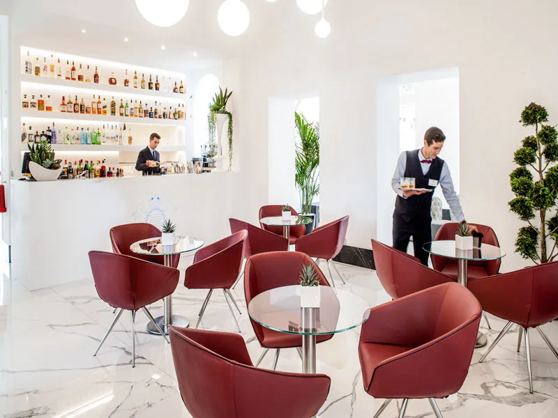 Bar interno moderno con poltrone rosse, tavolini in vetro e barman al bancone con bottiglie illuminate
