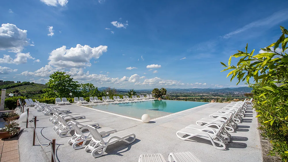 Ampia piscina a sfioro circondata da lettini bianchi con cielo azzurro e nuvole