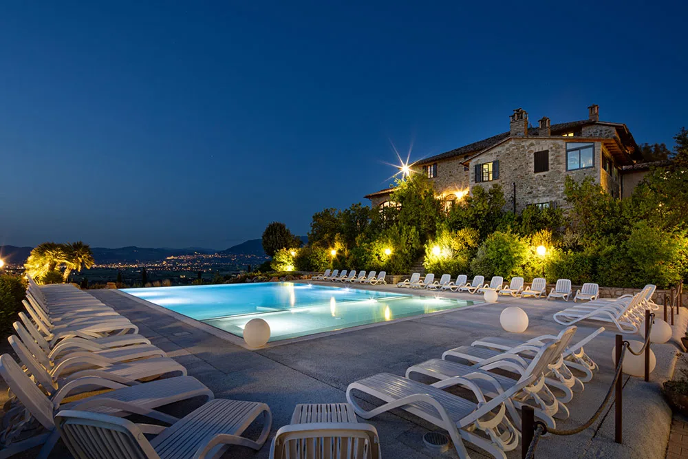 Piscina illuminata di notte con lettini bianchi e casale in pietra sullo sfondo sotto il cielo blu