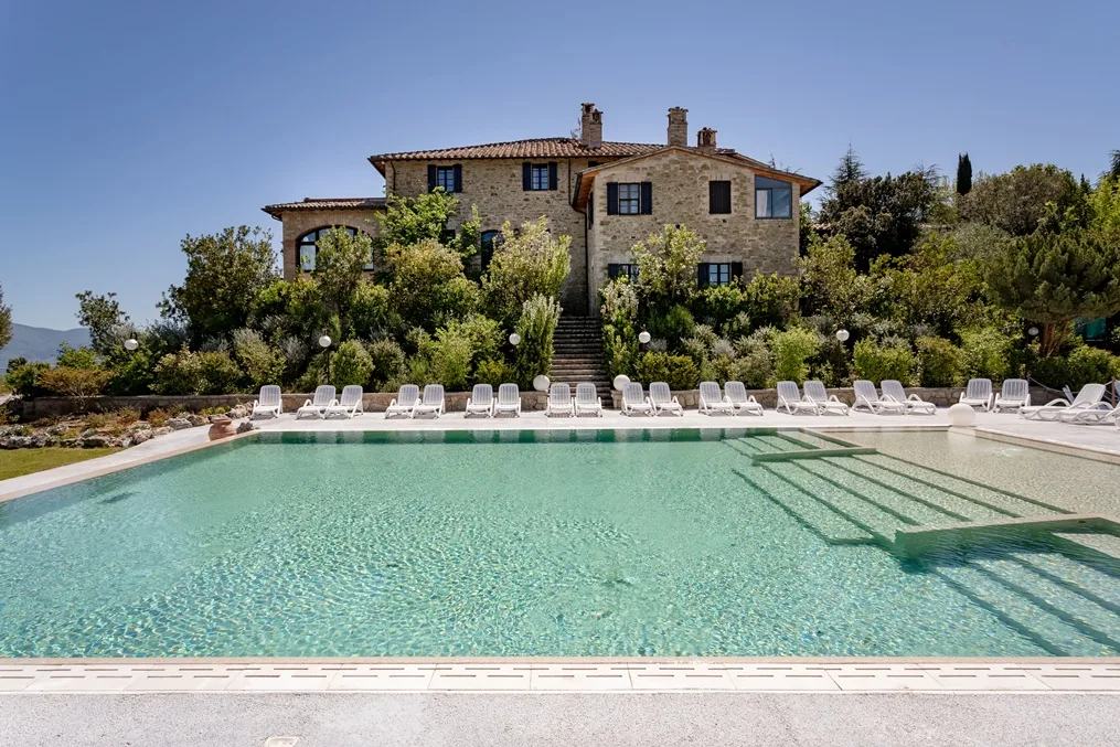 Piscina con acqua limpida e lettini bianchi davanti al casale in pietra circondato dal verde