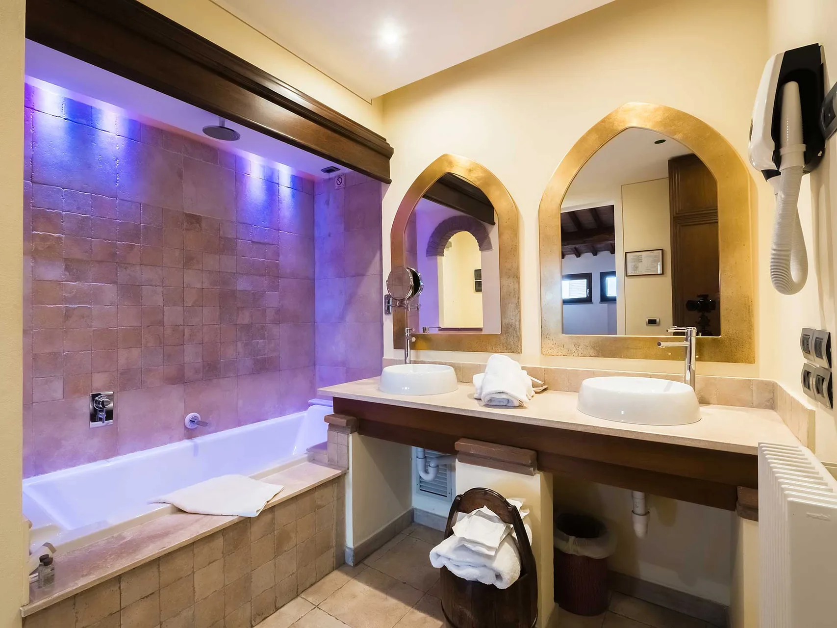 Bagno con vasca a doccia con illuminazione viola, doppio lavabo e specchi ad arco decorati