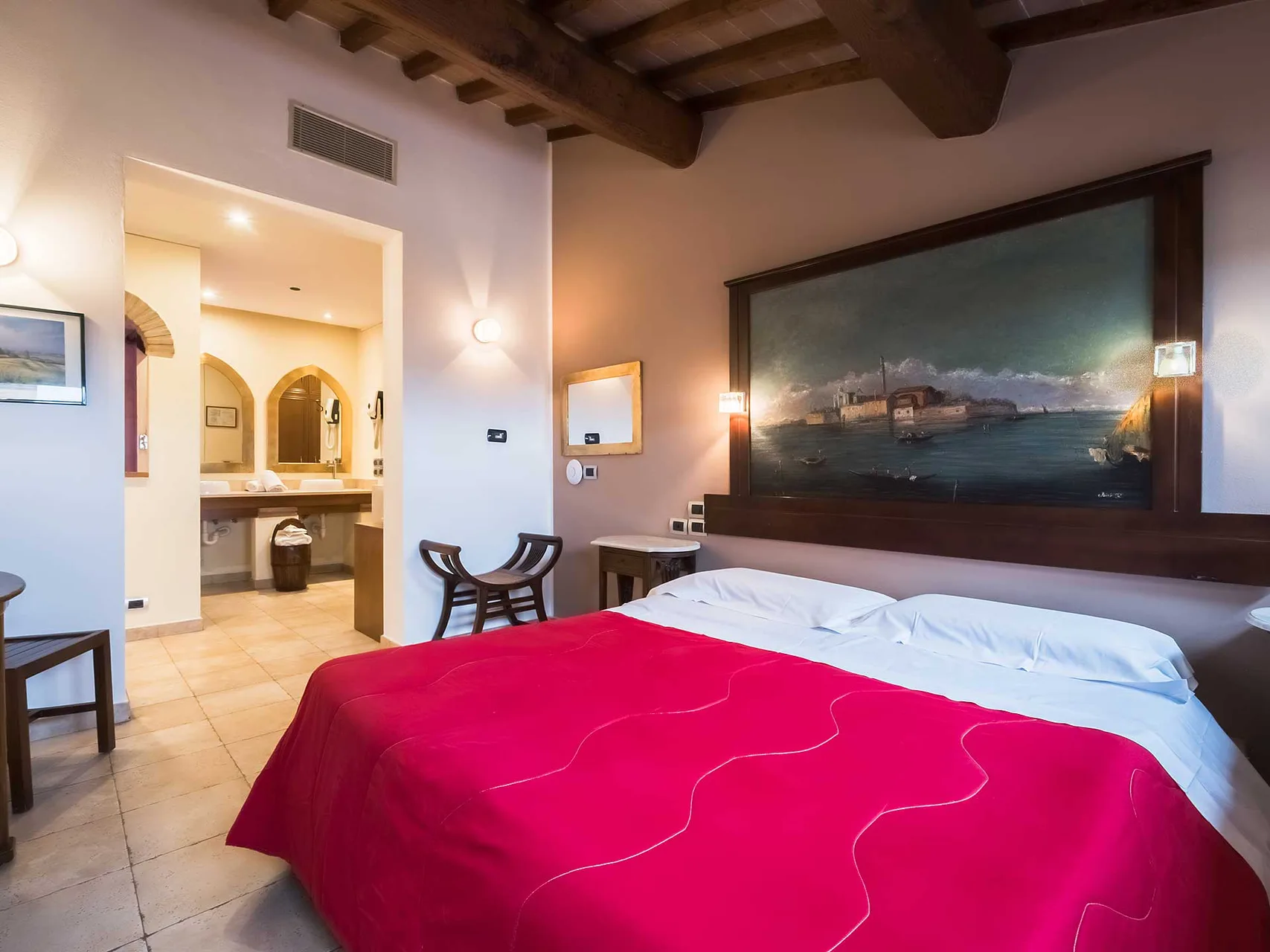 Camera matrimoniale con copriletto rosso, grande quadro paesaggistico e bagno open space sullo sfondo