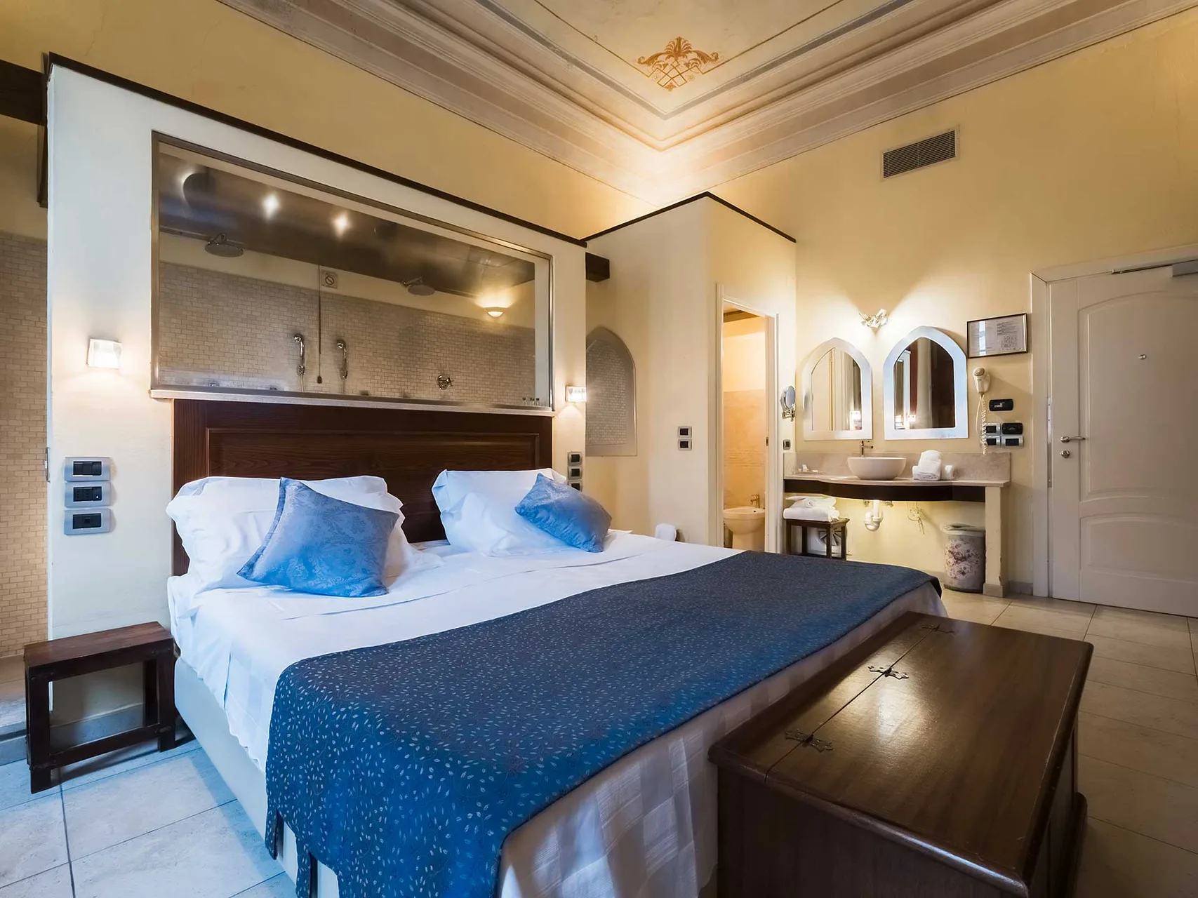 Camera elegante con soffitto affrescato, doccia a vista dietro il letto e zona lavabo integrata