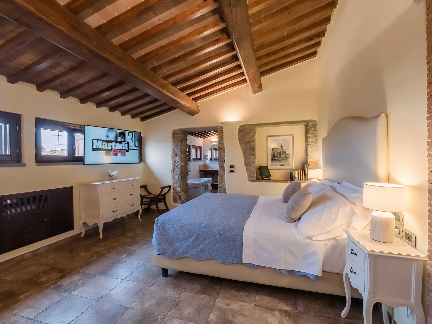 Camera matrimoniale con soffitto in travi di legno, TV a parete, dettagli in pietra e arredi bianchi