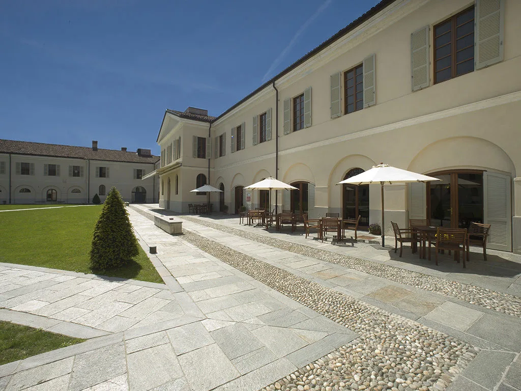 Cortile con pavimentazione in pietra e ciottoli, tavolini con ombrelloni bianchi accanto agli edifici color crema