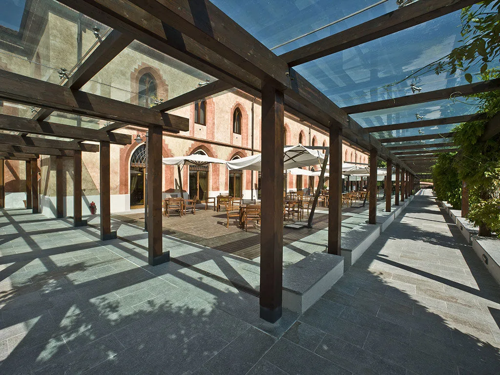Porticato moderno con travi in legno e copertura in vetro affacciato su area con tavoli e ombrelloni