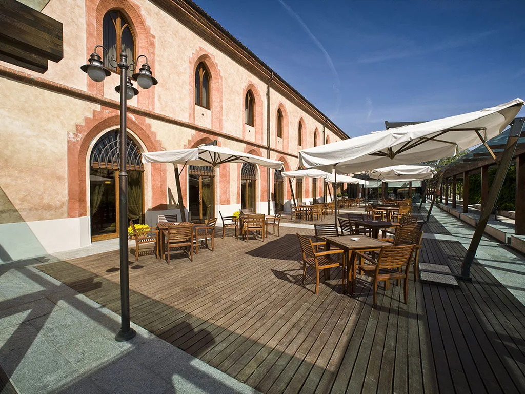 Terrazza esterna in legno con tavoli, sedie e ombrelloni bianchi lungo la facciata storica rosata