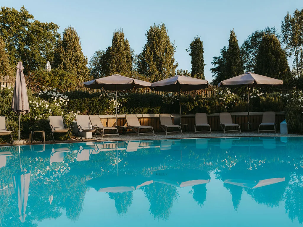 Piscina esterna con acqua turchese, lettini e ombrelloni riflessi nell'acqua tra alberi e giardino