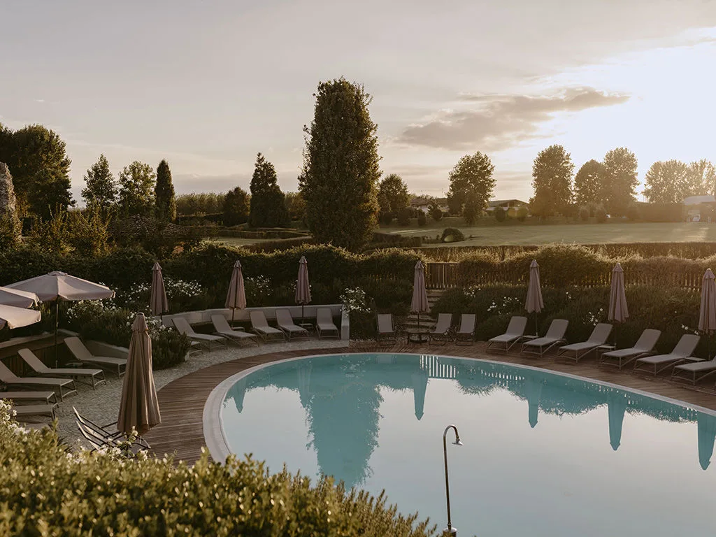 Piscina esterna con lettini e ombrelloni affacciata sul giardino e la campagna al tramonto