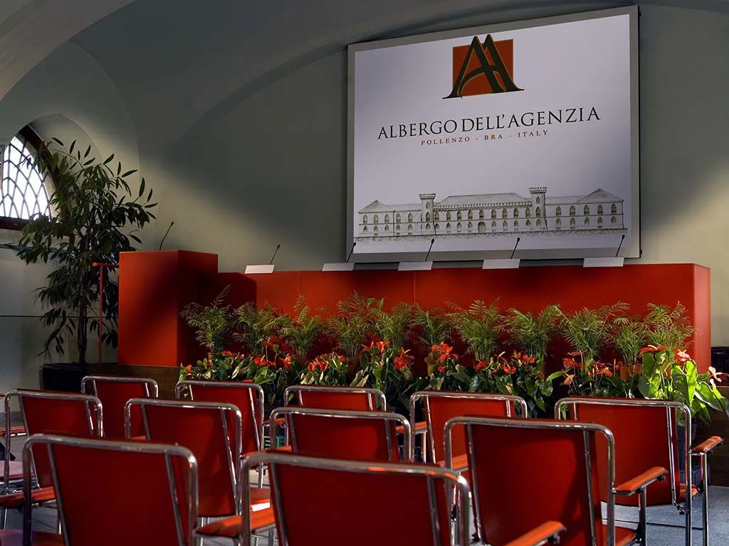 Sala conferenze con sedie rosse, palco, piante ornamentali e schermo con logo Albergo dell'Agenzia