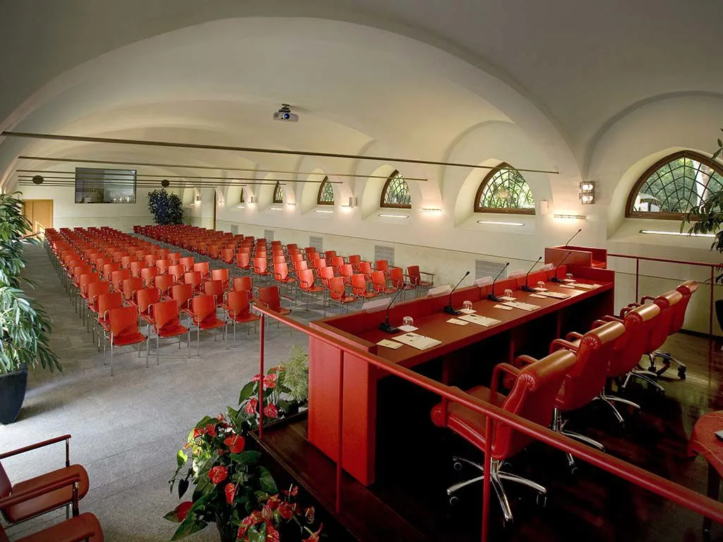 Ampia sala meeting con soffitto a volte, file di sedie rosse e tavolo relatori attrezzato