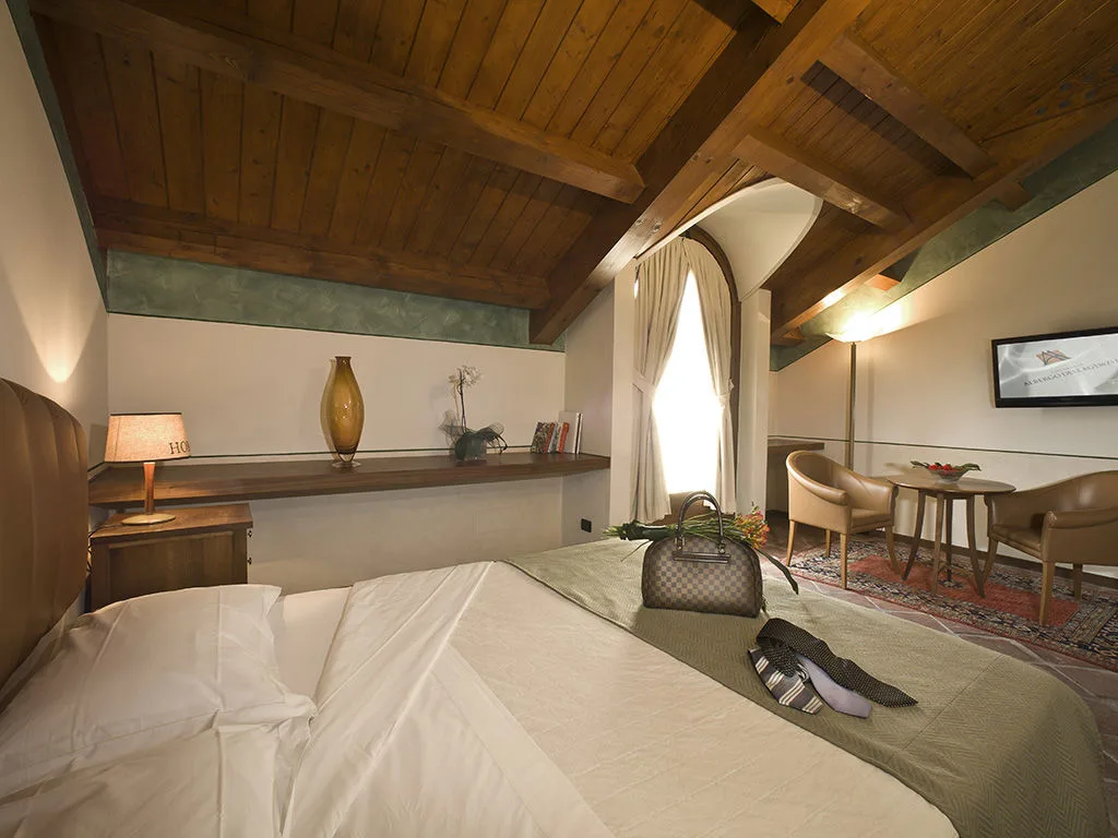Camera con soffitto in legno a vista, letto matrimoniale, area salotto e finestra ad arco
