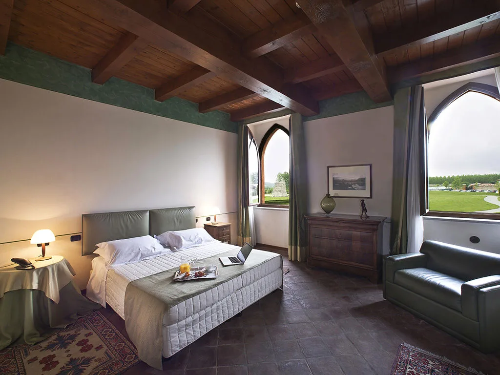 Camera con soffitto a travi in legno, letto matrimoniale, divano e finestre ad arco con vista sul giardino
