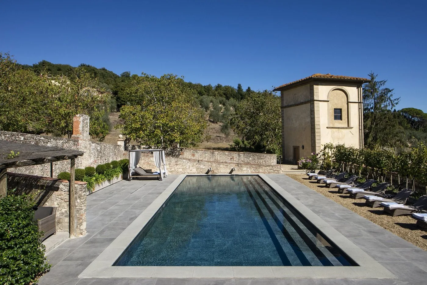 Piscina rettangolare con lettini e antica torre in pietra sullo sfondo delle colline