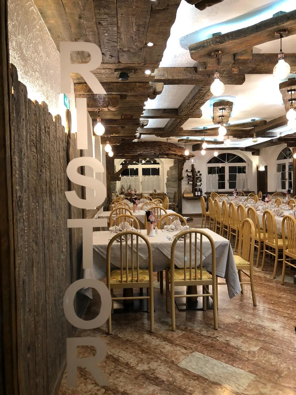 Sala ristorante rustica con travi in legno a vista, tavoli apparecchiati e sedie chiare
