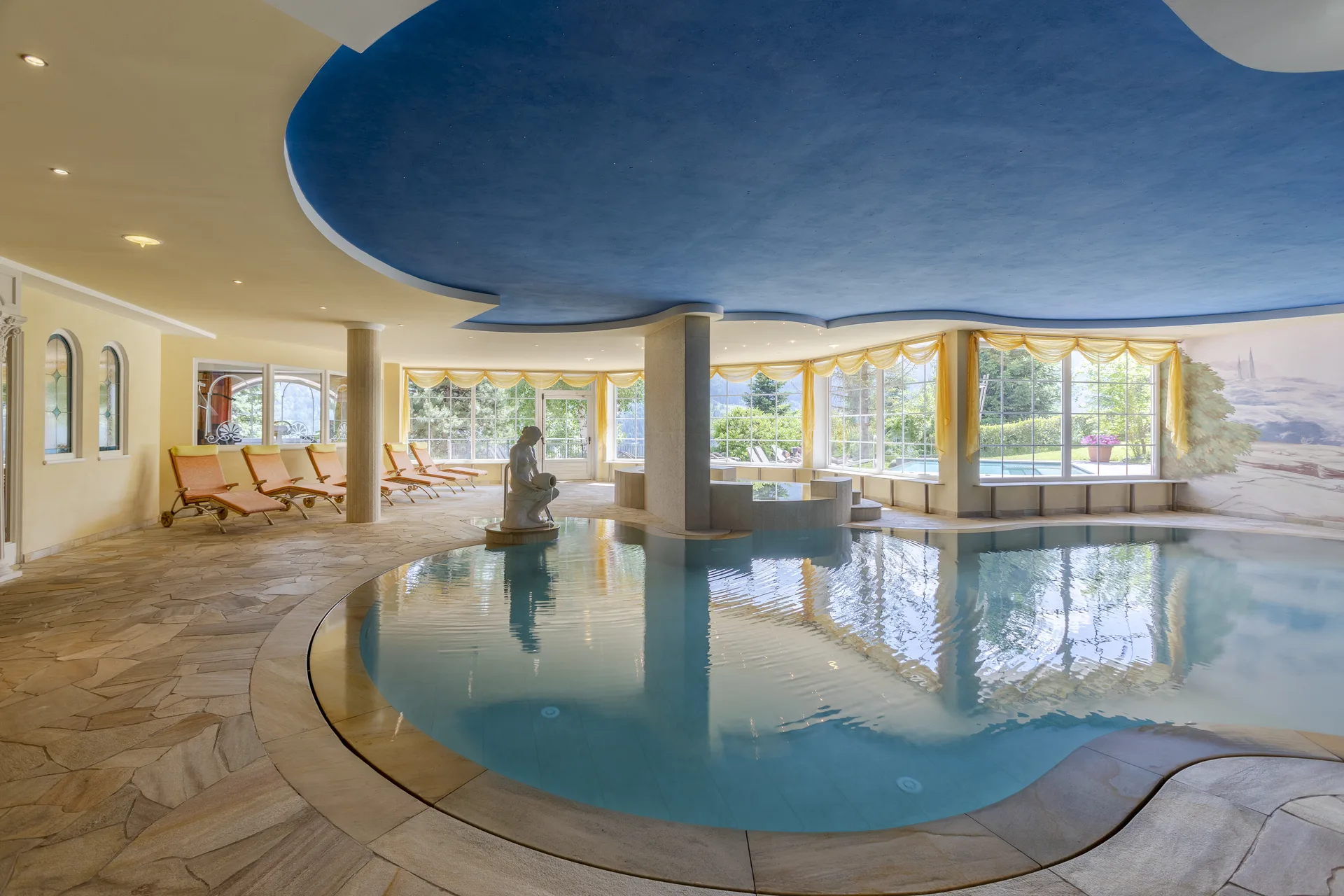 Piscina interna con soffitto blu, lettini arancioni e ampie vetrate sul giardino