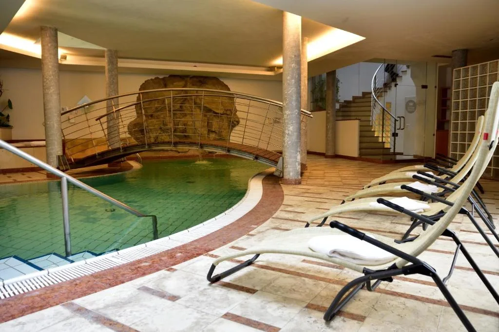 Area piscina coperta con lettini, ponte in legno e parete rocciosa decorativa
