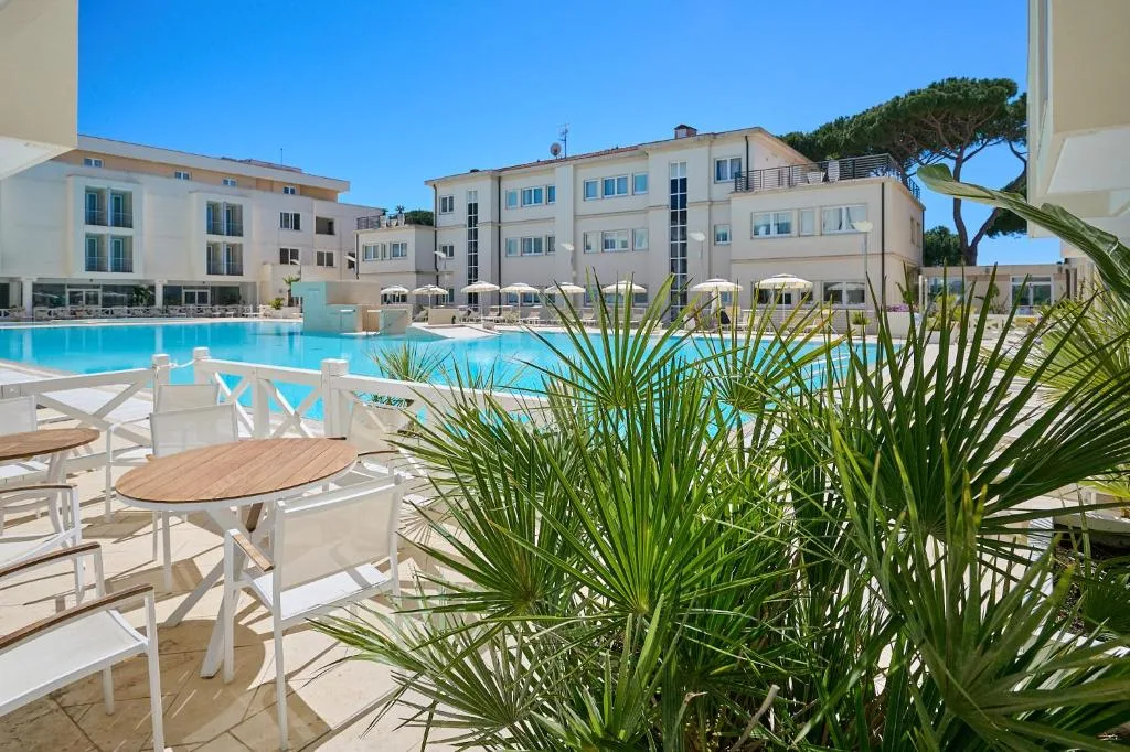 Area lounge esterna con tavolini e sedie bianche vicino alla piscina e alla vegetazione