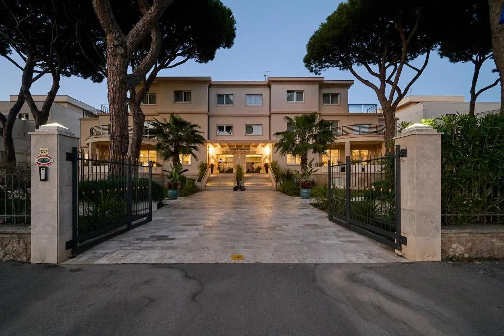 Ingresso dell'hotel al tramonto con cancello, palme e viale illuminato