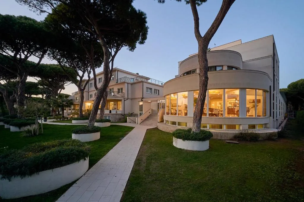 Esterno dell'hotel con giardino curato, pini e sale con ampie vetrate illuminate al crepuscolo