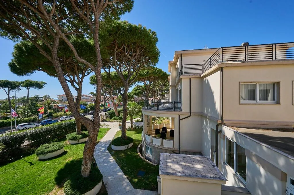 Vista del giardino con pini marittimi e terrazza dell'hotel sotto il cielo azzurro
