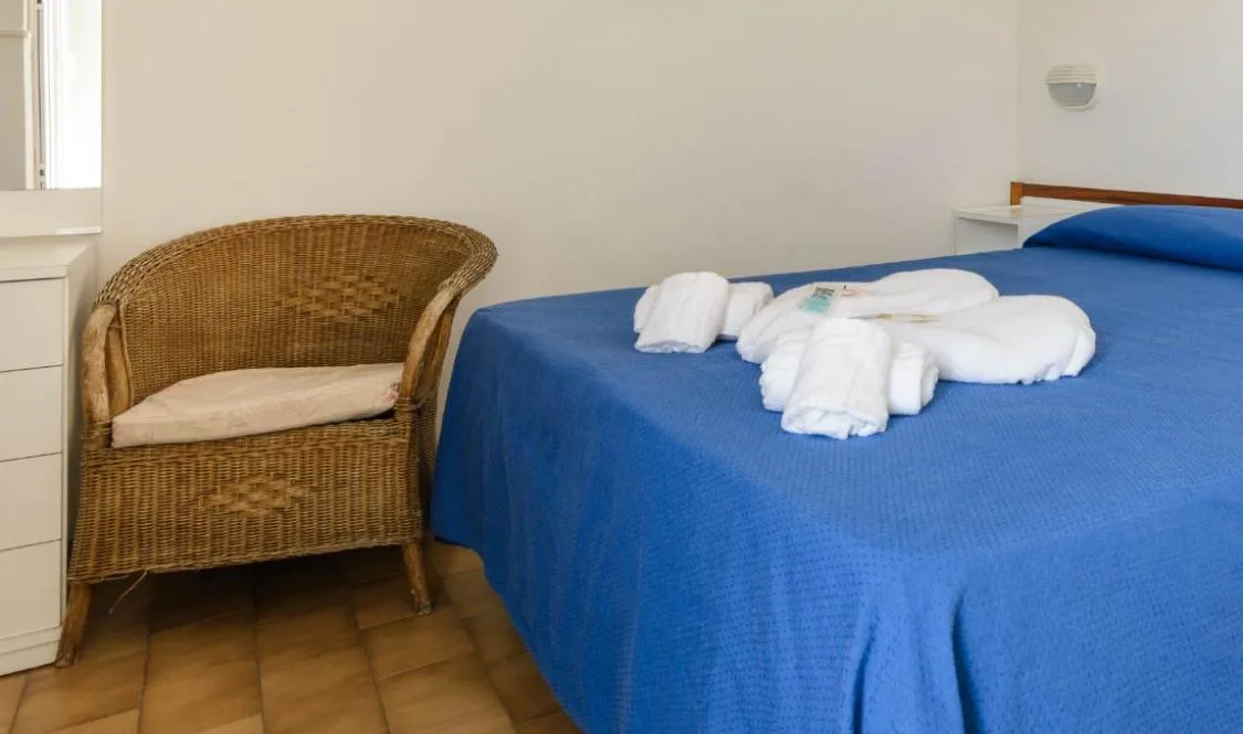 Dettaglio camera con letto matrimoniale, copriletto blu, poltrona in vimini e asciugamani