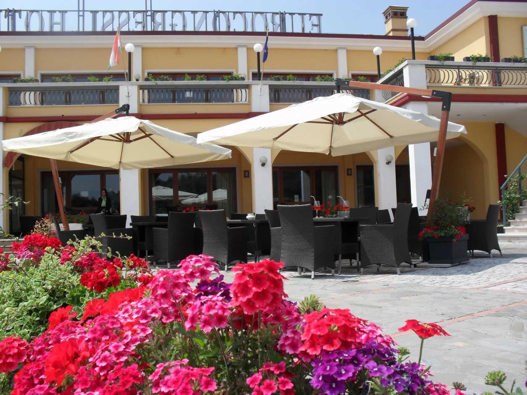 Terrazza esterna con ombrelloni bianchi, tavoli e aiuole fiorite davanti alla facciata dell'hotel
