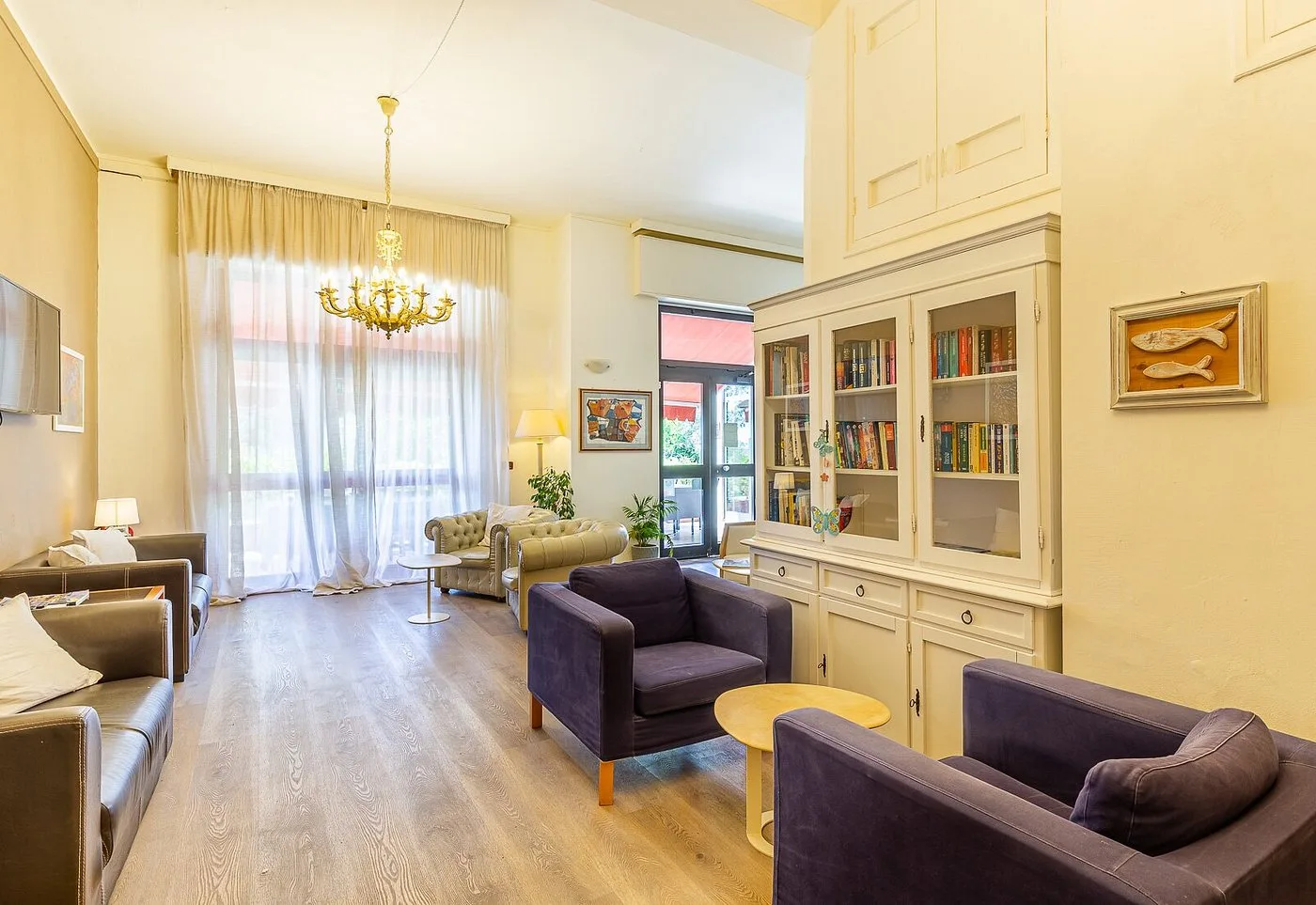 Salotto luminoso con poltrone, libreria bianca, lampadario dorato e ampie tende