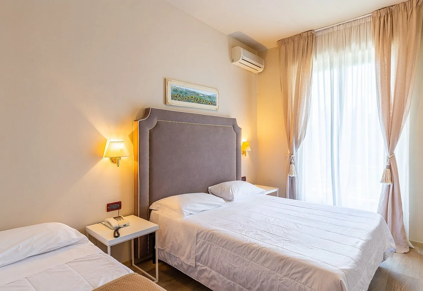 Camera con letto matrimoniale, letto singolo, testiera imbottita e ampia finestra con tende