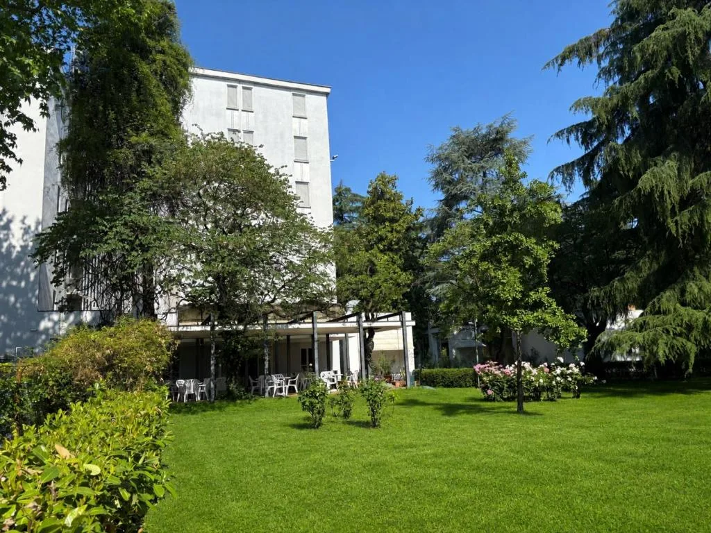 Esterno dell'hotel con giardino verde, alberi e terrazza con tavolini