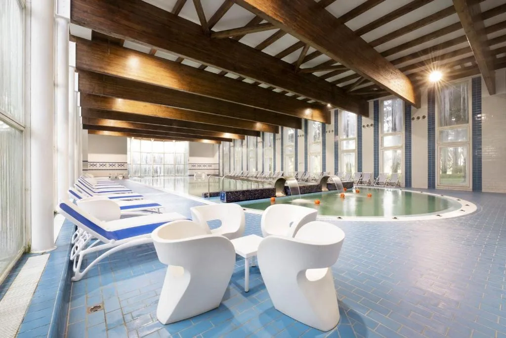 Piscina interna con lettini, poltrone bianche di design e soffitto con travi in legno