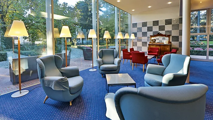 Hall dell'hotel con poltrone azzurre, lampade da terra e moquette blu vicino a grandi vetrate