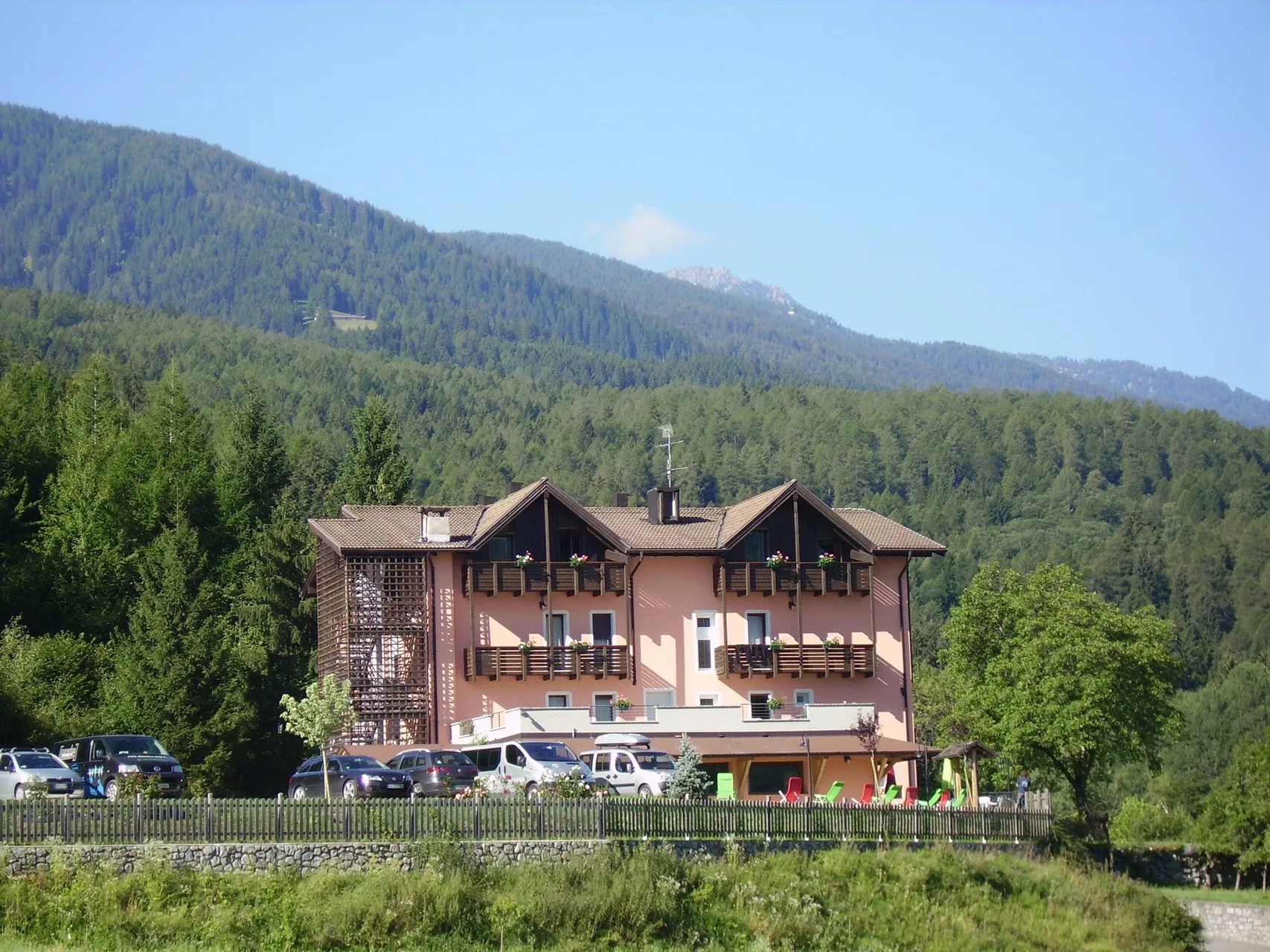 Vista estiva dell'hotel immerso nel bosco con montagne sullo sfondo e auto parcheggiate