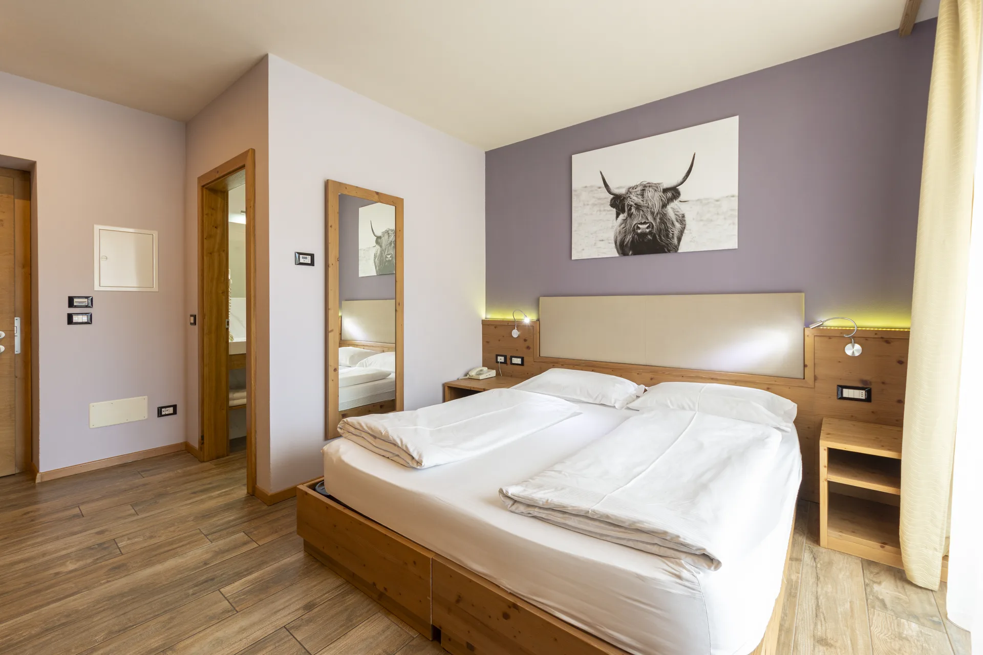 Camera matrimoniale con parete lilla, arredo in legno e quadro con mucca highland
