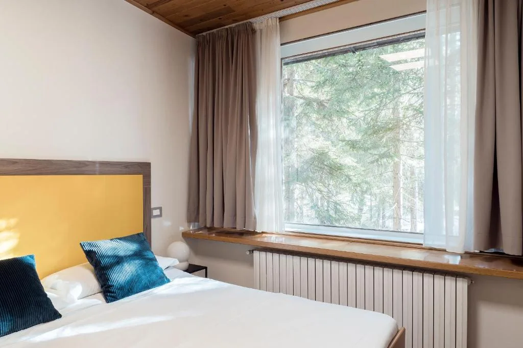 Camera da letto con testiera gialla, cuscini blu e finestra con vista sul bosco
