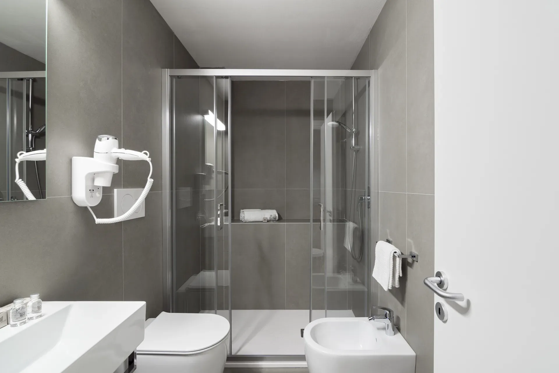 Bagno moderno grigio con doccia in vetro, WC, bidet e lavabo con asciugacapelli