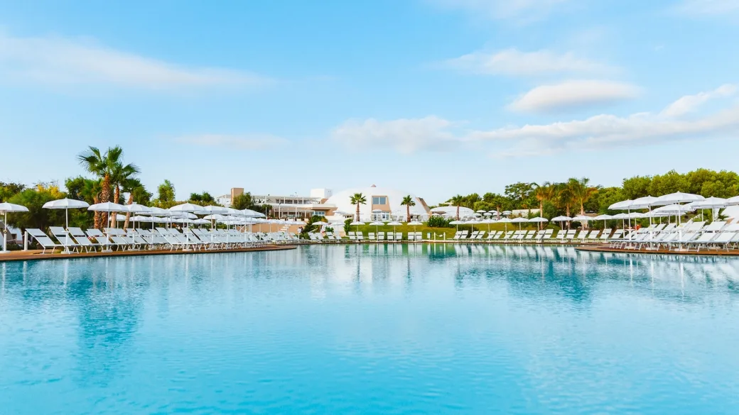 Ampia piscina con lettini e ombrelloni bianchi e vista sulla struttura del resort