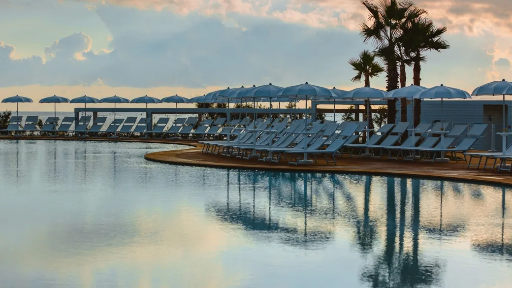 Piscina con lettini azzurri, ombrelloni e palme al tramonto vista mare