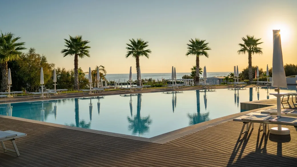 Piscina con palme e lettini sul deck in legno al tramonto vista mare