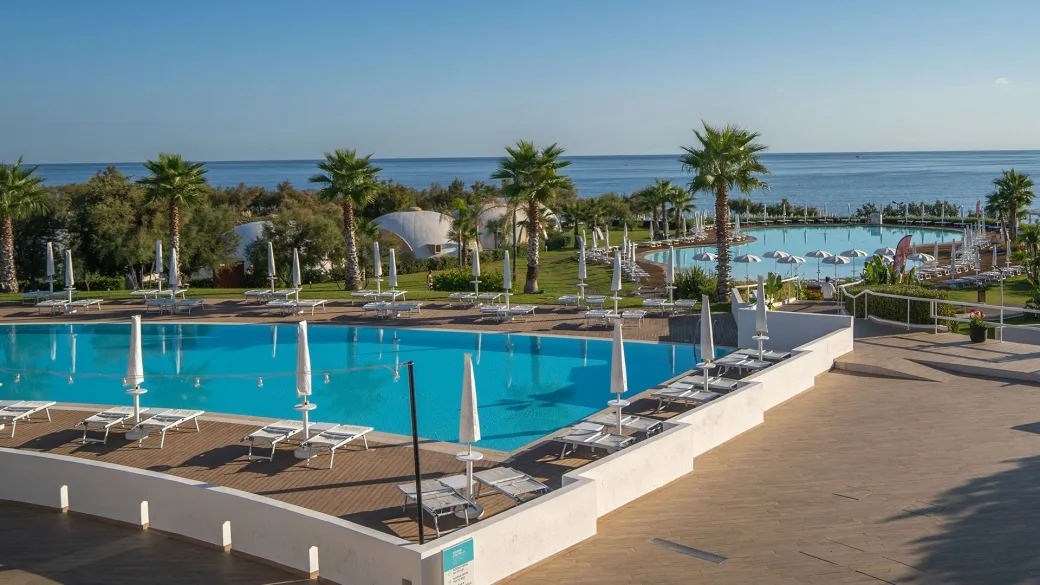 Vista su due piscine con lettini, ombrelloni e palme affacciate sul mare