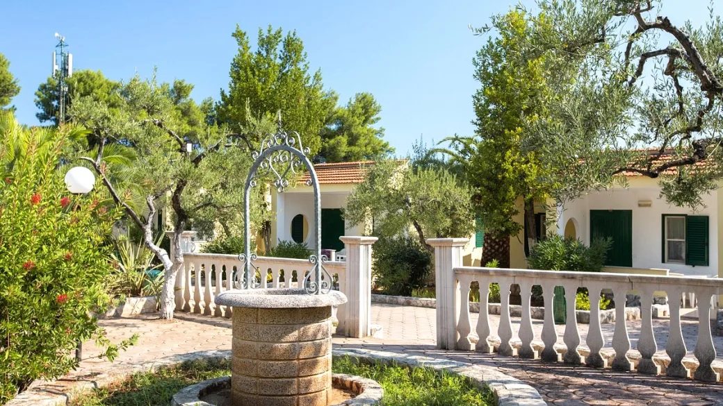 Giardino con pozzo decorativo in pietra, balaustra bianca e villette tra ulivi e oleandri