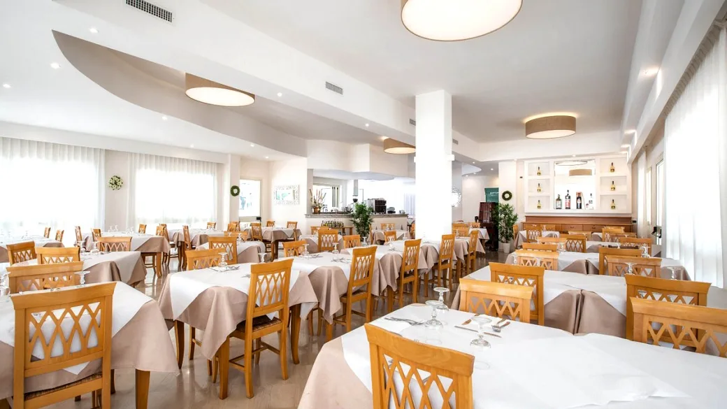 Ampia sala ristorante moderna con tavoli bianchi, sedie in legno e grandi finestre luminose