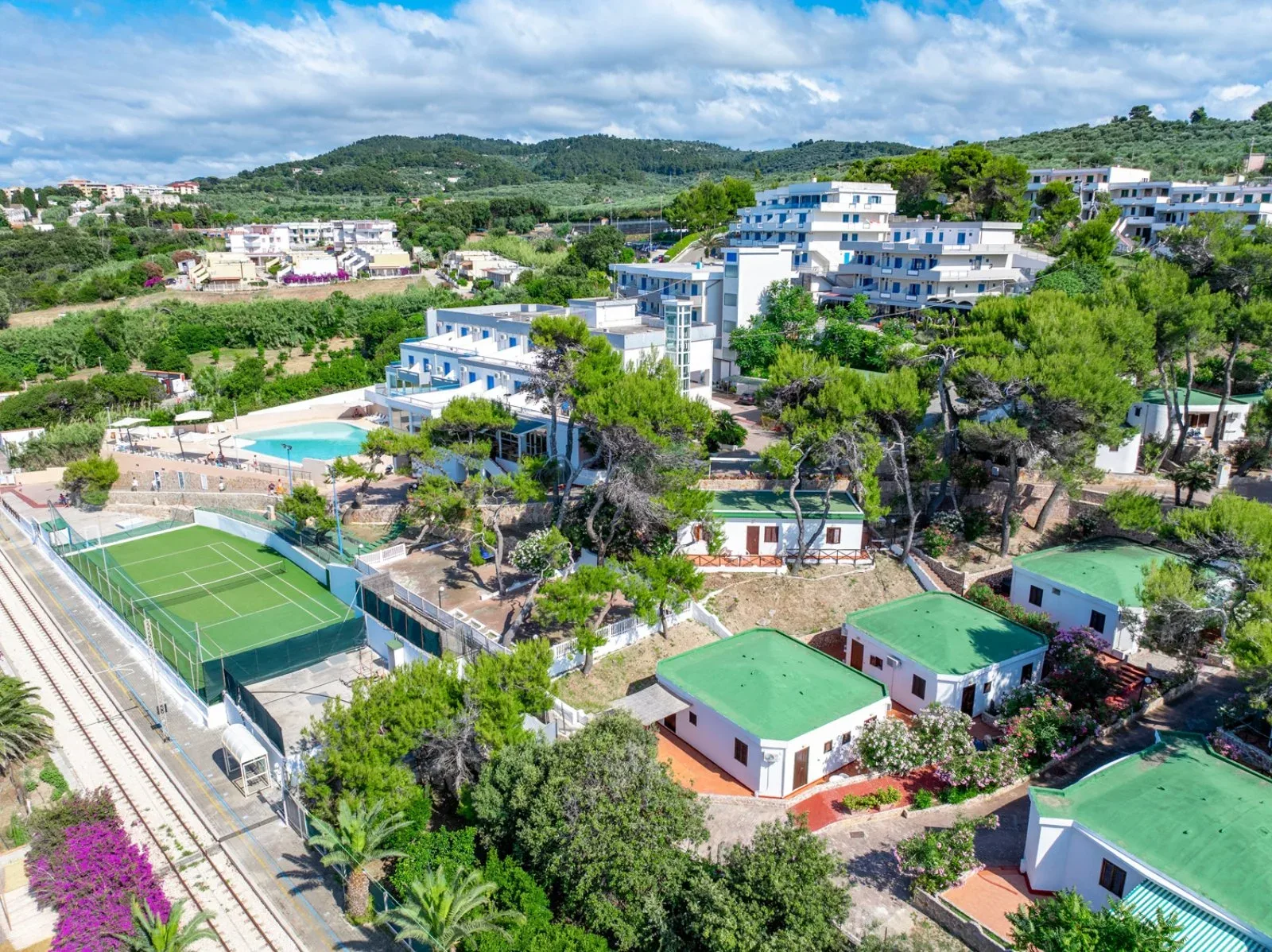 Vista aerea del resort tra il verde con piscina, campo da tennis e bungalow con tetto verde