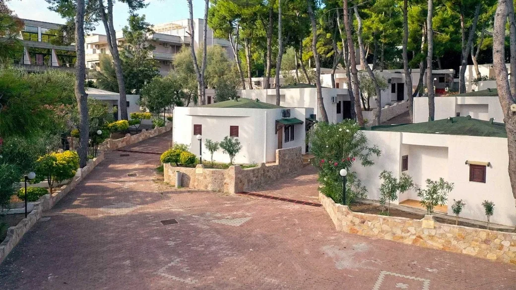 Viale tra bungalow bianchi immersi nel verde con pini e piante mediterranee