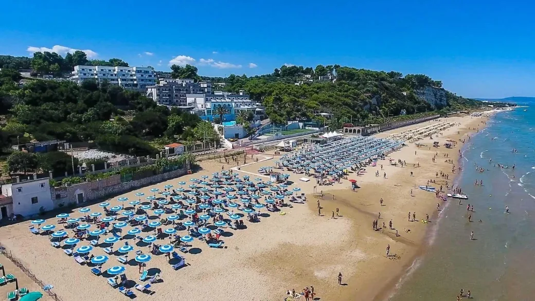 Vista aerea della spiaggia con file di ombrelloni, bagnanti e struttura dell'hotel sulla collina