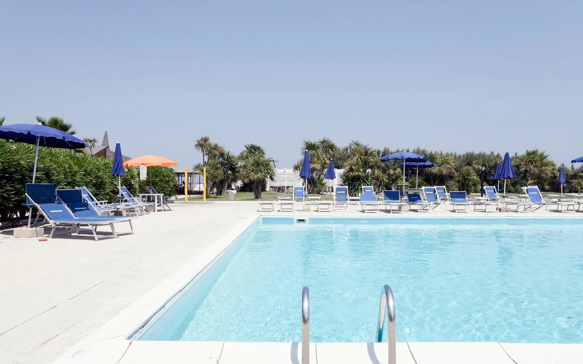 Piscina con acqua cristallina, lettini blu e ombrelloni colorati con palme sullo sfondo