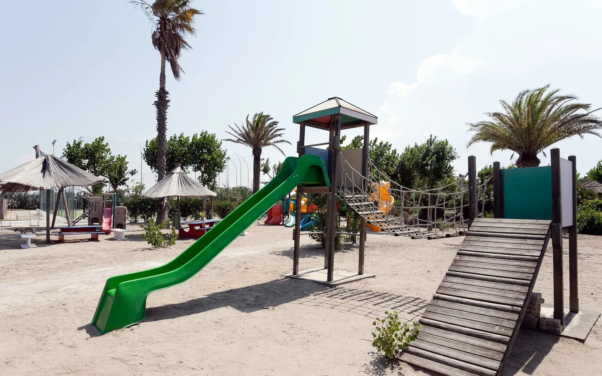 Parco giochi per bambini con scivolo verde, ponti in legno e palme