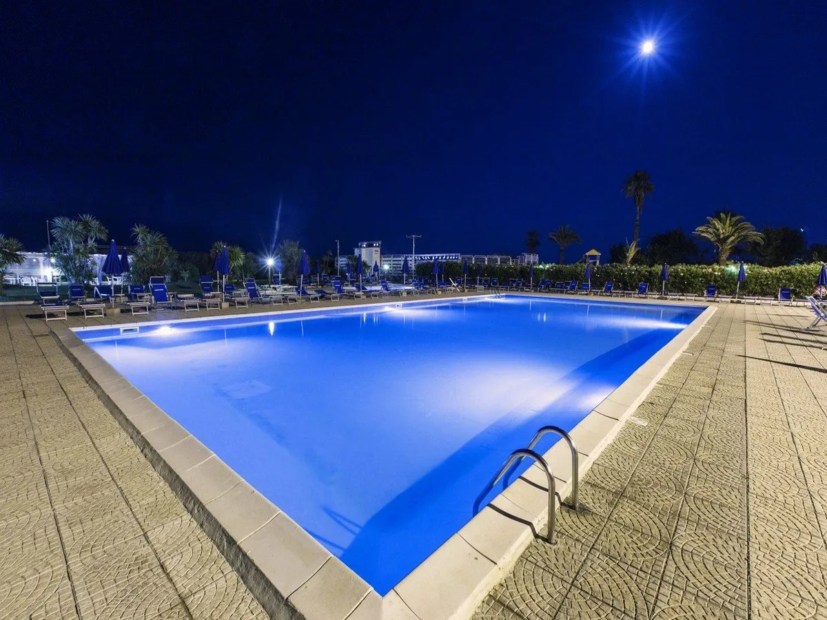 Piscina illuminata di notte con lettini, ombrelloni e luna nel cielo