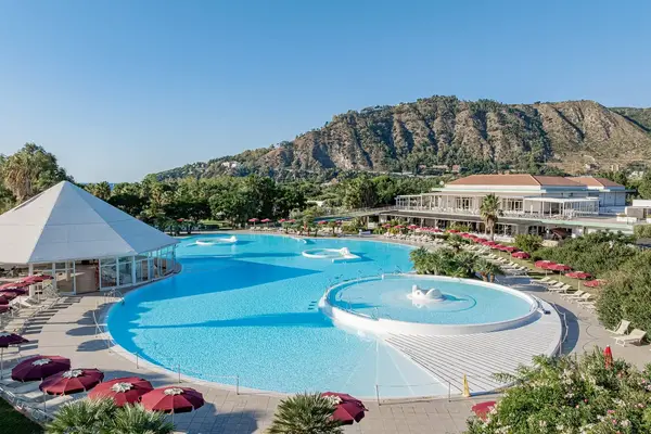 Ampia piscina del resort con gazebo bianco, ombrelloni bordeaux e montagna sullo sfondo
