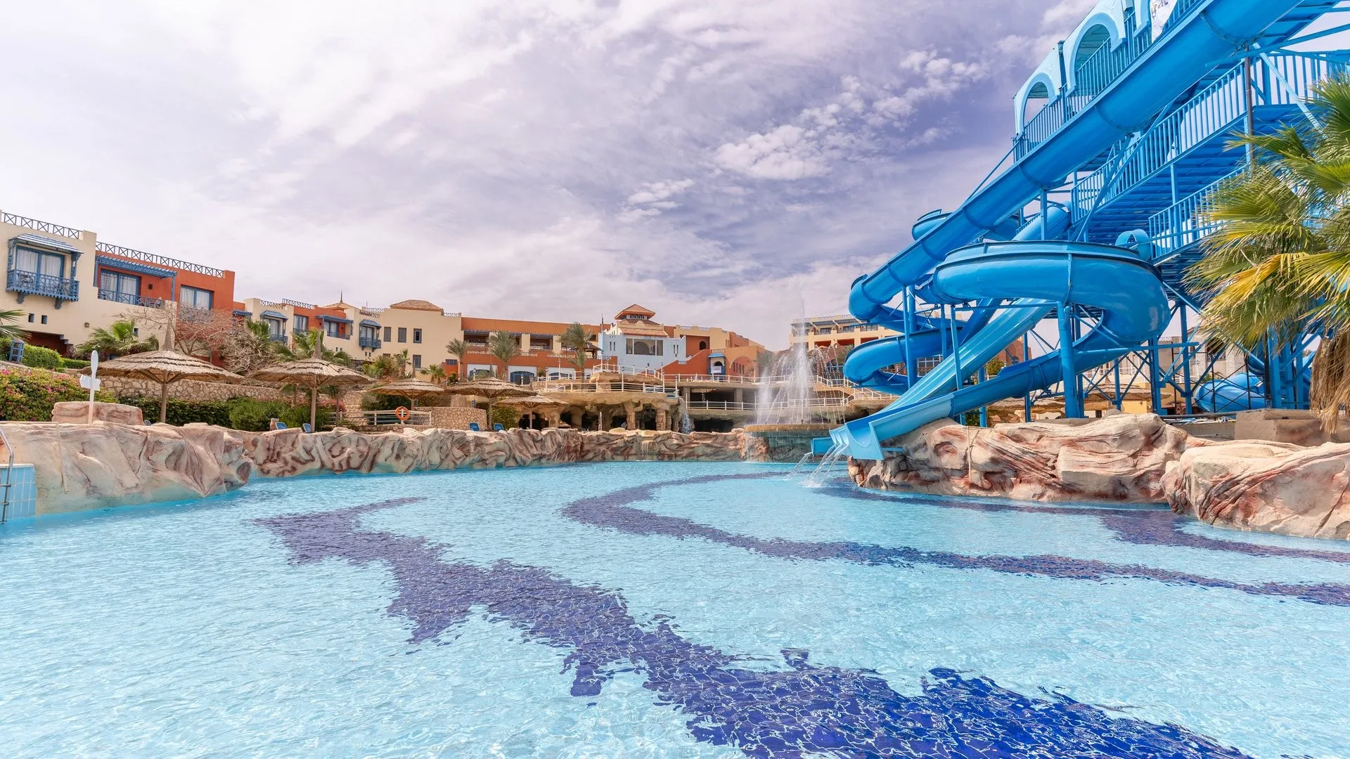 Piscina con scivoli acquatici blu, fontana e ombrelloni in paglia circondata dagli edifici del resort